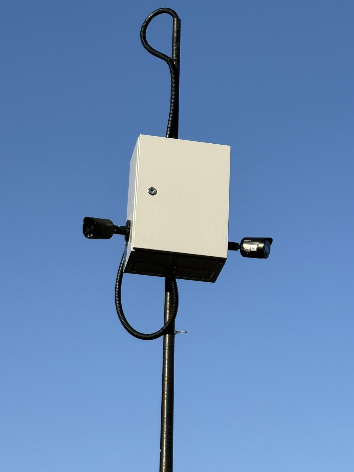Słup z zamontowaną białą skrzynką i dwiema kamerami monitoringu na tle błękitnego nieba.