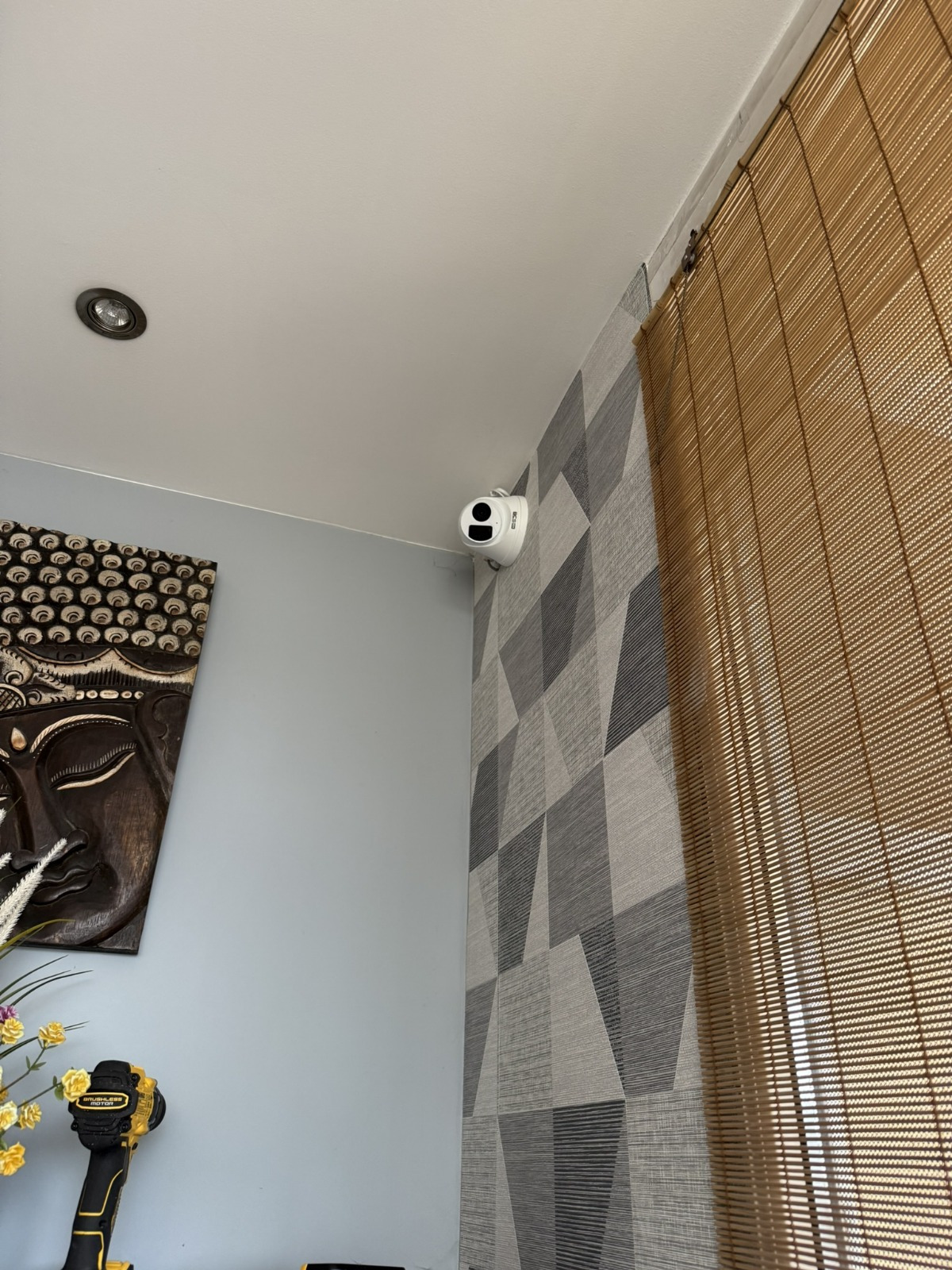 Biała kamera monitoringu zamontowana w rogu pokoju na ścianie z geometryczną tapetą, obok bambusowa roleta i dekoracyjna maska na ścianie. W tle widoczna lampa sufitowa.