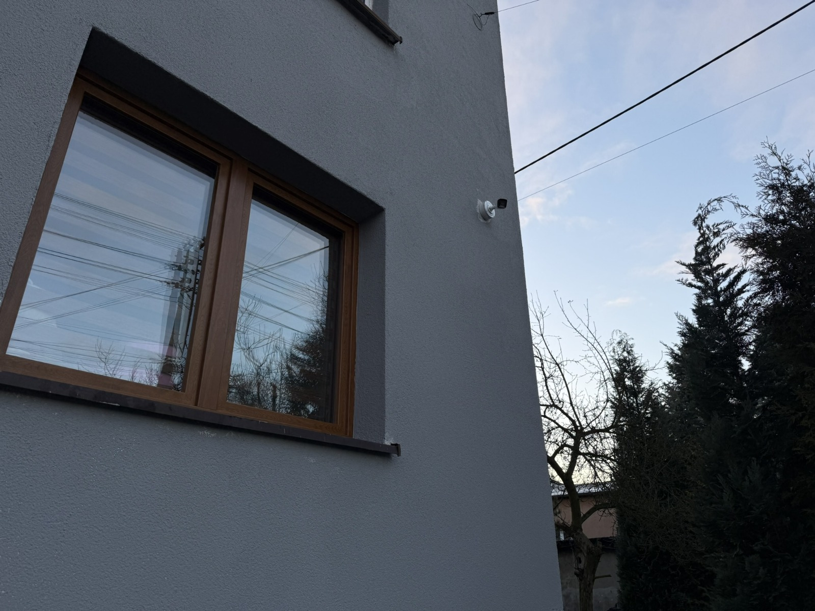 Zewnętrzna kamera monitoringu na szarej ścianie budynku, widoczna z boku, z oknem z brązową ramą i drzewami w tle.