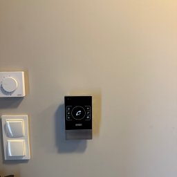 NEXTGEN DAWID TOMASZEWSKI - Nowoczesny, czarny panel domofonu STech zamontowany na jasnej ścianie, obok włącznika światła i termostatu. Minimalistyczny design, widoczne ikony funkcji.