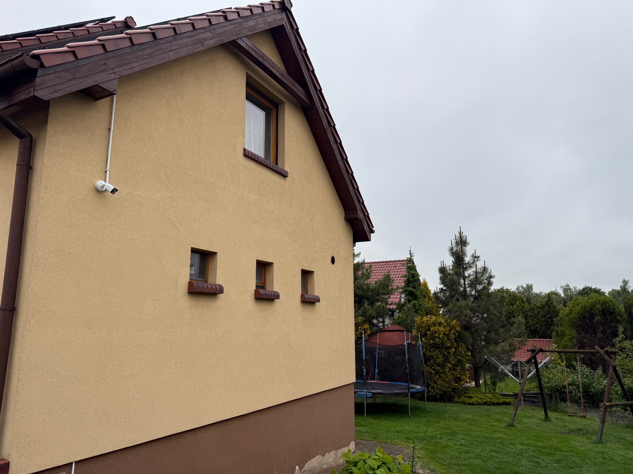 Kamera monitoringu zamontowana na elewacji domu z beżowym tynkiem, widok z boku, na tle zielonego ogrodu z trampoliną i huśtawką.
