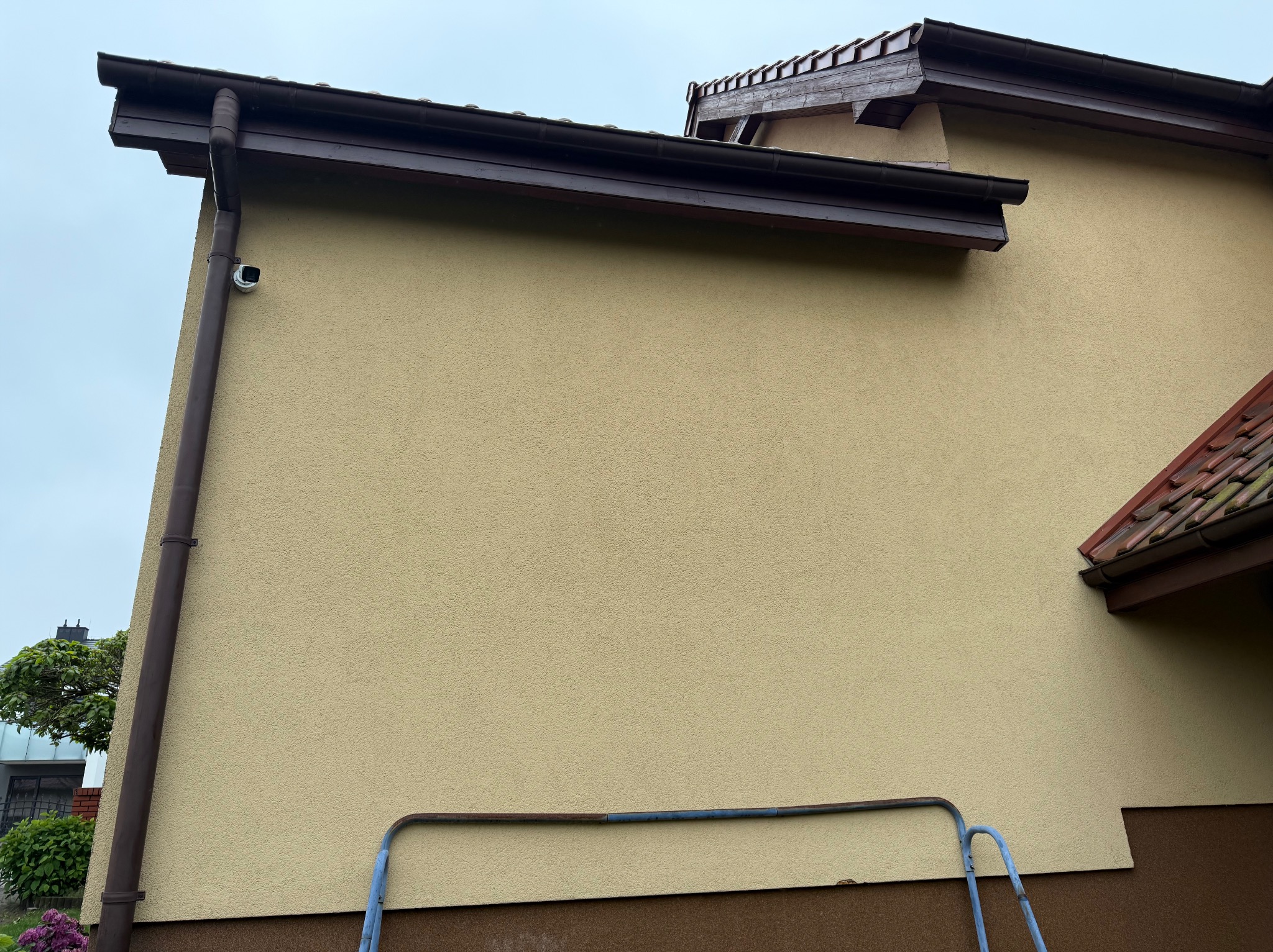 Kamera monitoringu zamontowana na elewacji domu z brązowymi rynnami. Na dole fragment drabiny. Ujęcie z dołu, pochmurne niebo.