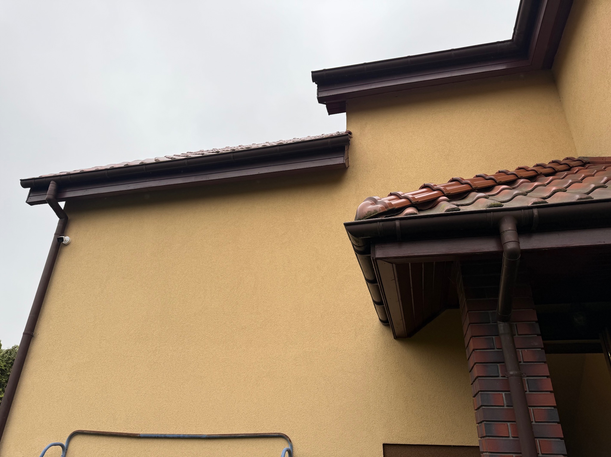 Kamera na elewacji domu z brązowymi rynnami i fragmentem dachu z dachówką. Ujęcie z dołu, pochmurne niebo.