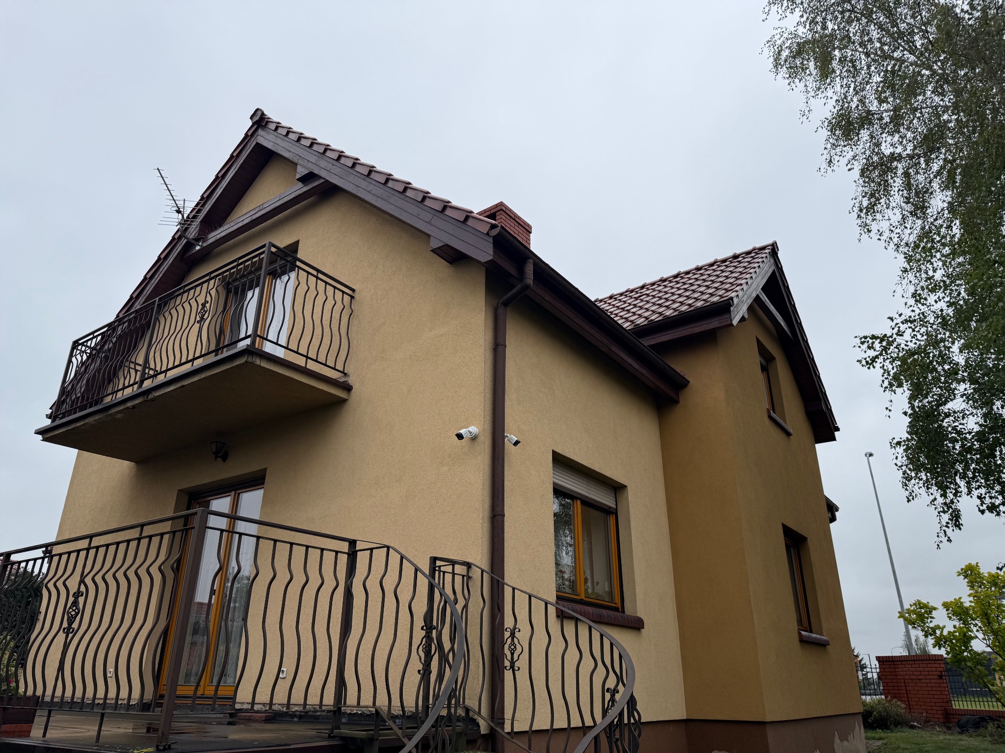 Dwukondygnacyjny dom z balkonami i monitoringiem: dwie białe kamery na elewacji. Brązowa dachówka, okna i balustrady. Antena na dachu. Ujęcie z dołu, pochmurne niebo.
