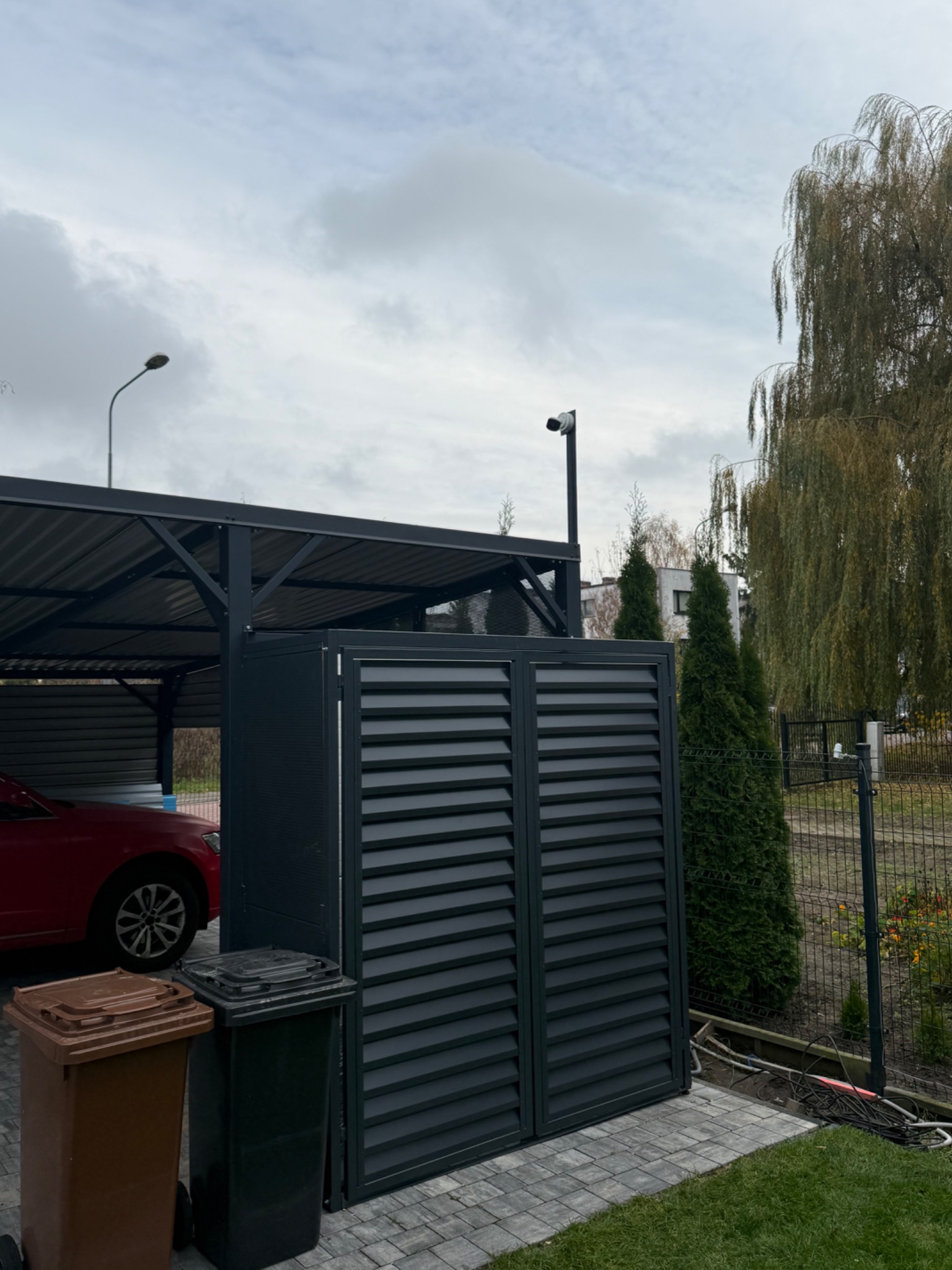 Zewnętrzna kamera monitoringu na słupku, widoczna nad zabudową śmietnikową z lamelami, obok wiaty garażowej i drzewa płaczącej wierzby. Szara kostka brukowa.