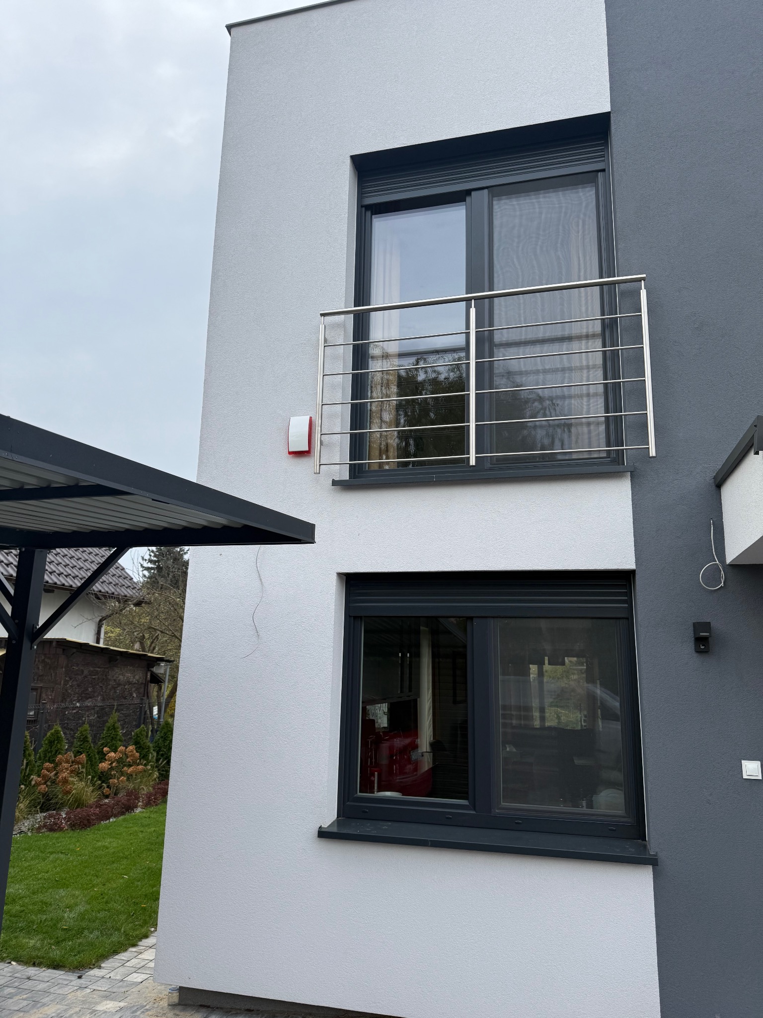 Dwukondygnacyjny dom z szarą elewacją, balkonem z metalową balustradą i zamontowanym alarmem na ścianie. Widoczne okna z roletami zewnętrznymi i fragment ogrodu.
