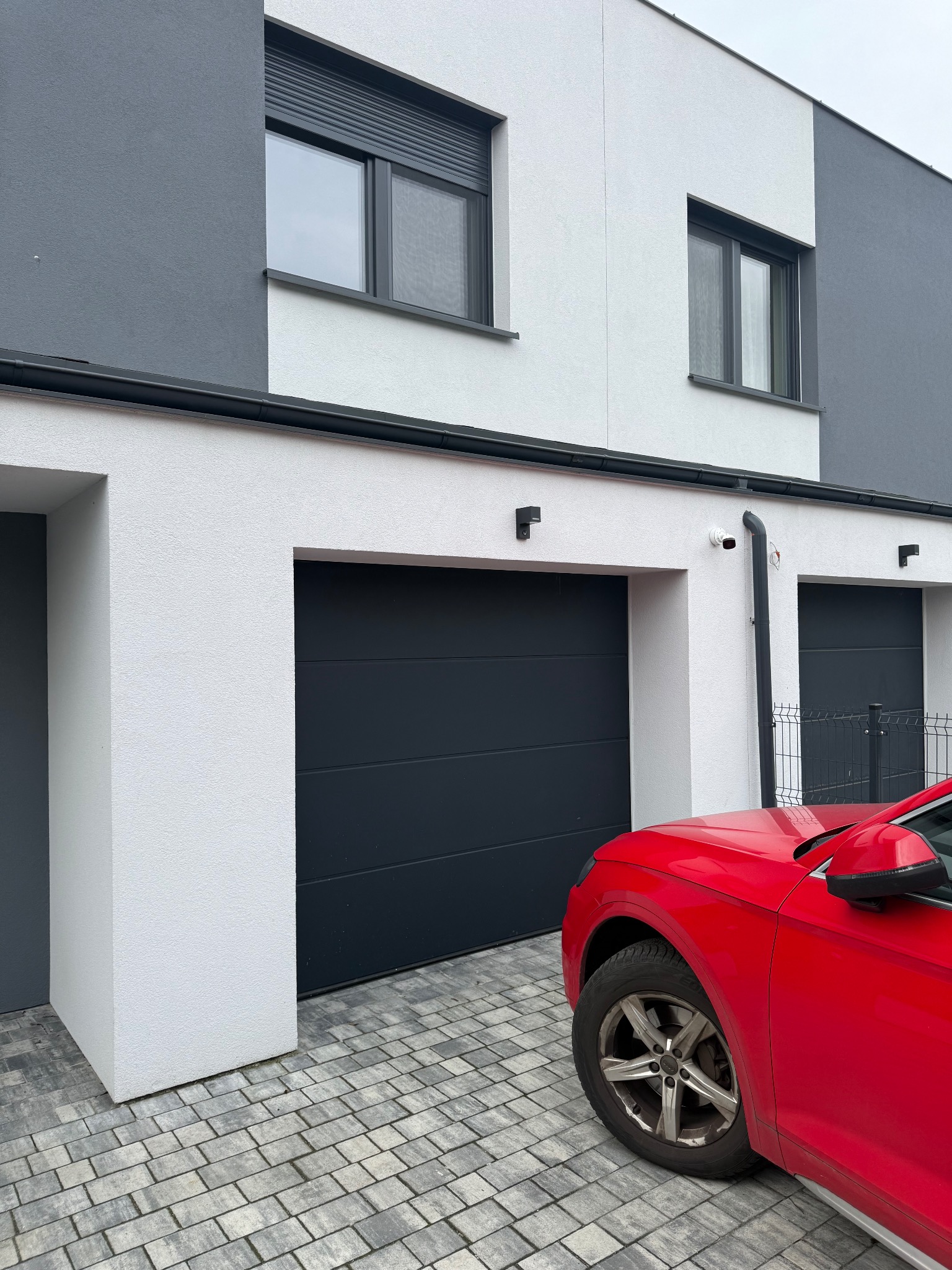 Nowoczesny dom z szarą elewacją, czarnymi bramami garażowymi i widoczną kamerą monitoringu. Czerwony samochód częściowo zasłania widok na posesję.