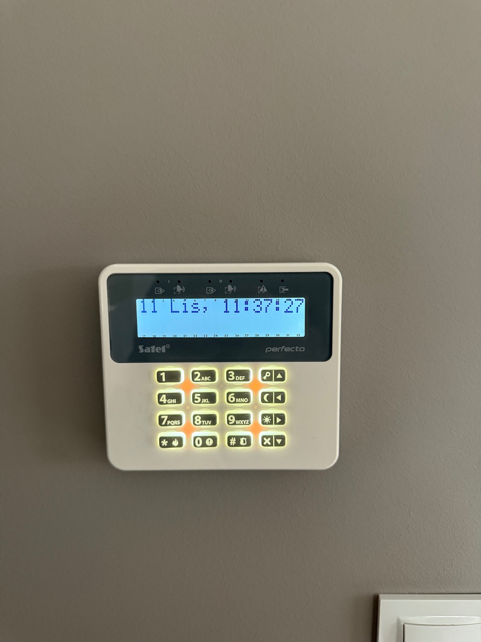 Panel sterowania alarmem Satel Perfecta na szarej ścianie. Wyświetlacz LCD pokazuje datę i godzinę. Podświetlane przyciski numeryczne i funkcyjne. Widoczny fragment gniazdka.