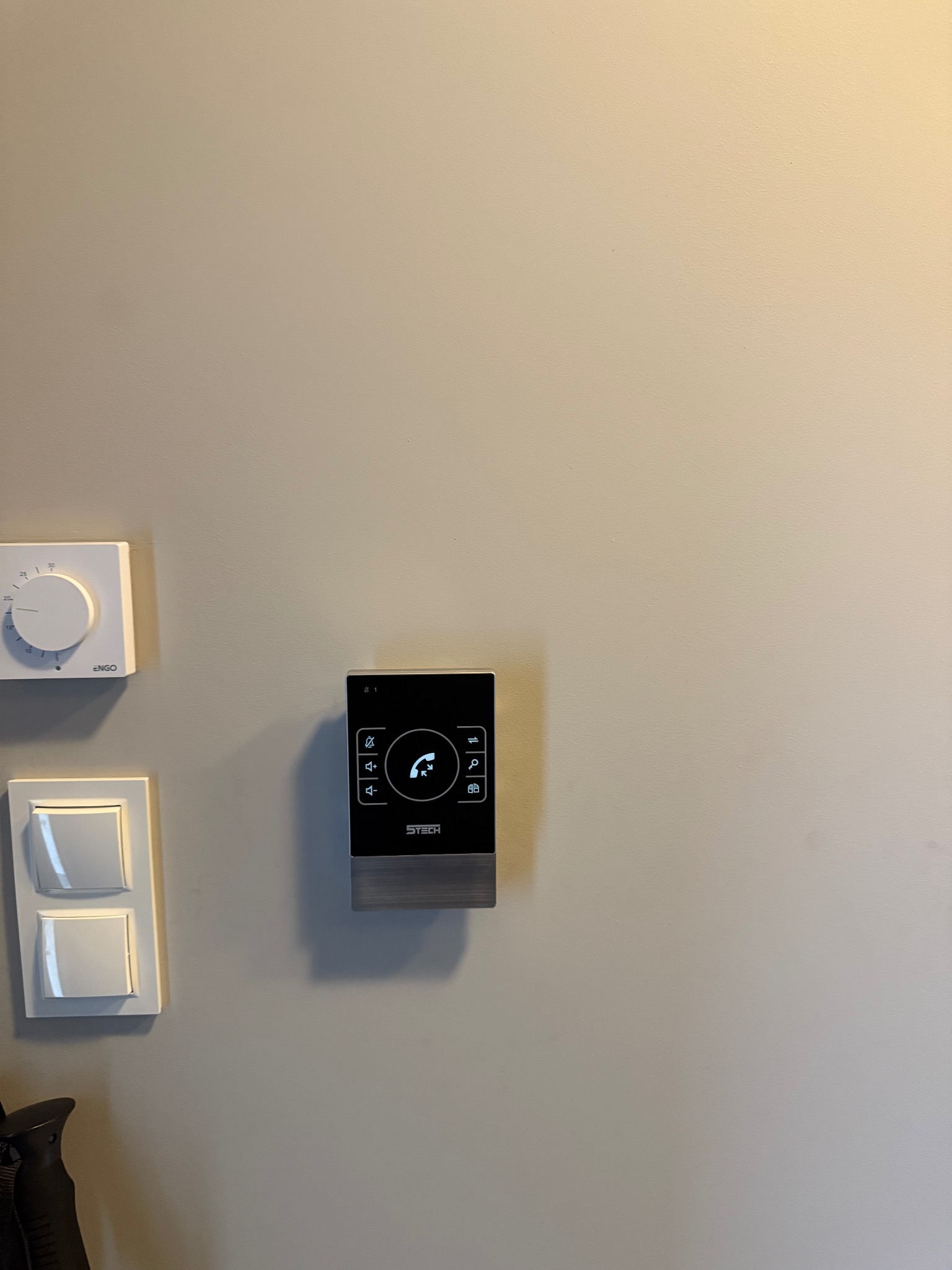 Nowoczesny, czarny panel domofonu STech zamontowany na jasnej ścianie, obok włącznika światła i termostatu. Minimalistyczny design, widoczne ikony funkcji.