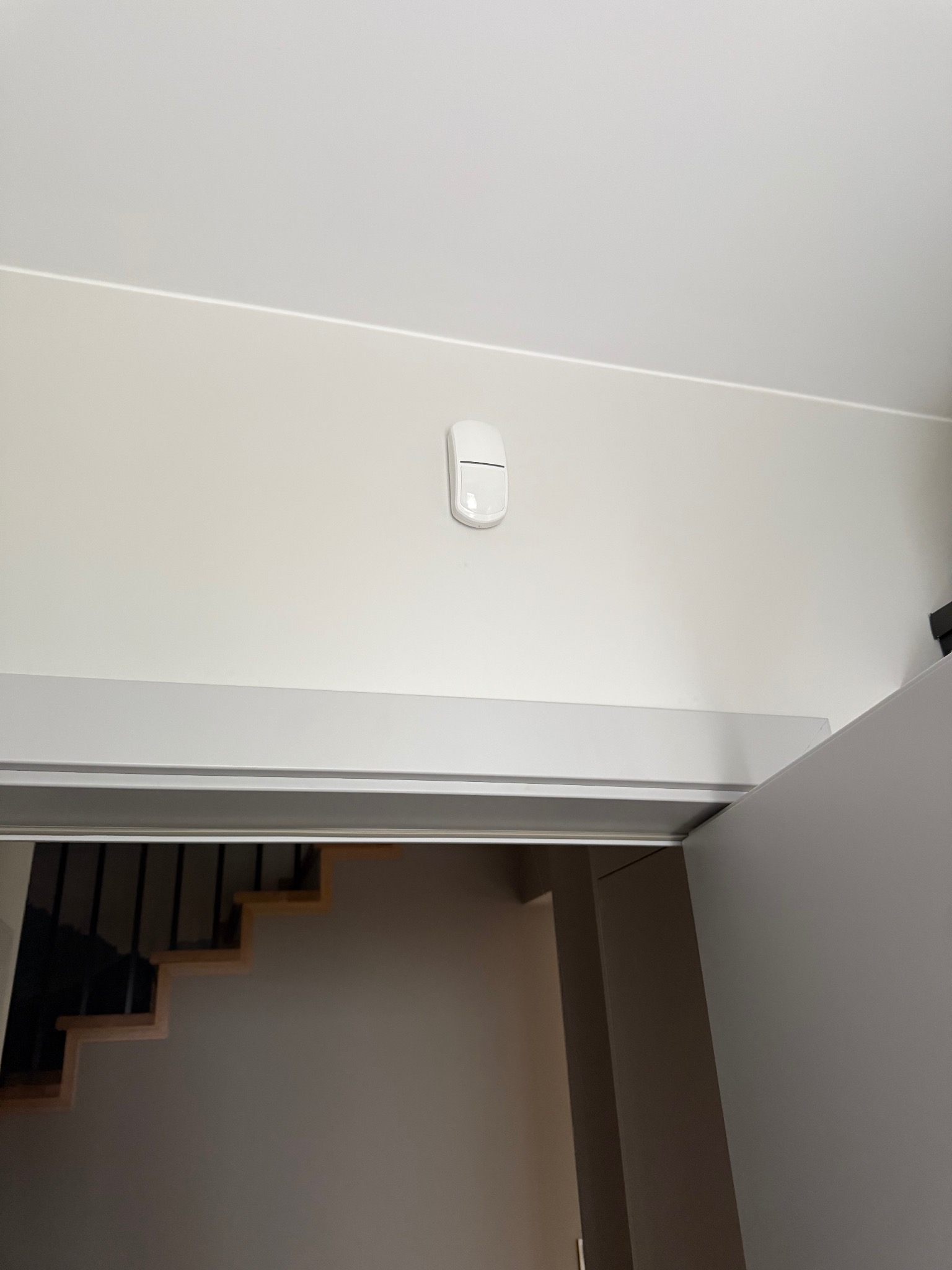 Biały czujnik alarmowy zamontowany na ścianie w korytarzu, widoczne schody w tle. Minimalistyczny design, neutralna kolorystyka.