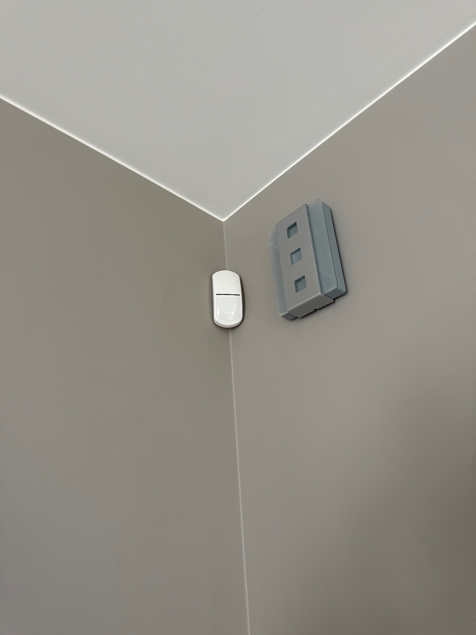 Zamontowany czujnik ruchu i moduł alarmowy na szarej ścianie w rogu pomieszczenia. Widok z dołu, oświetlenie naturalne.