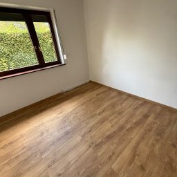 EPOXY BUD Kamil Tetlak - Pusty pokój po remoncie: drewnopodobna podłoga, białe ściany, okno z widokiem na zieleń. Świeżo wykończone wnętrze z widocznymi rurkami grzewczymi przy podłodze.