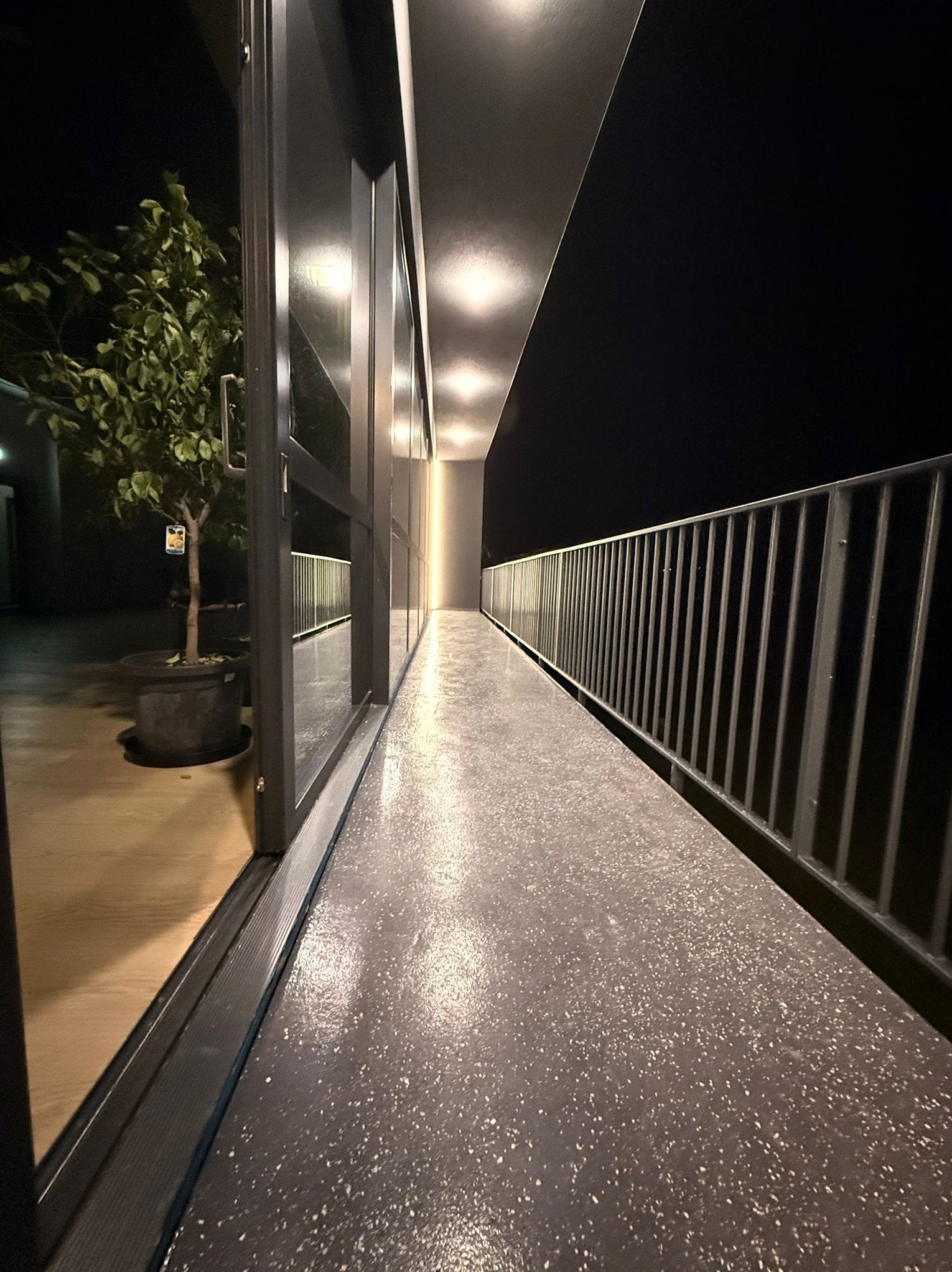 Balkon z szarą posadzką żywiczną, metalową balustradą i widokiem na drzewo w donicy, oświetlony punktowymi lampami sufitowymi, nocne ujęcie.