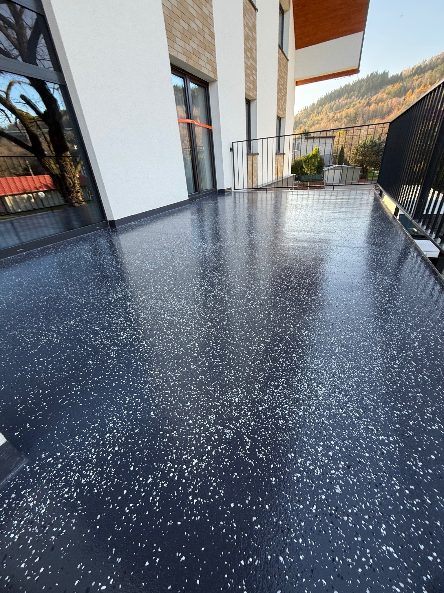 Błyszcząca, ciemna posadzka żywiczna z białymi drobinkami na balkonie nowoczesnego domu z widokiem na zalesione wzgórza w tle.