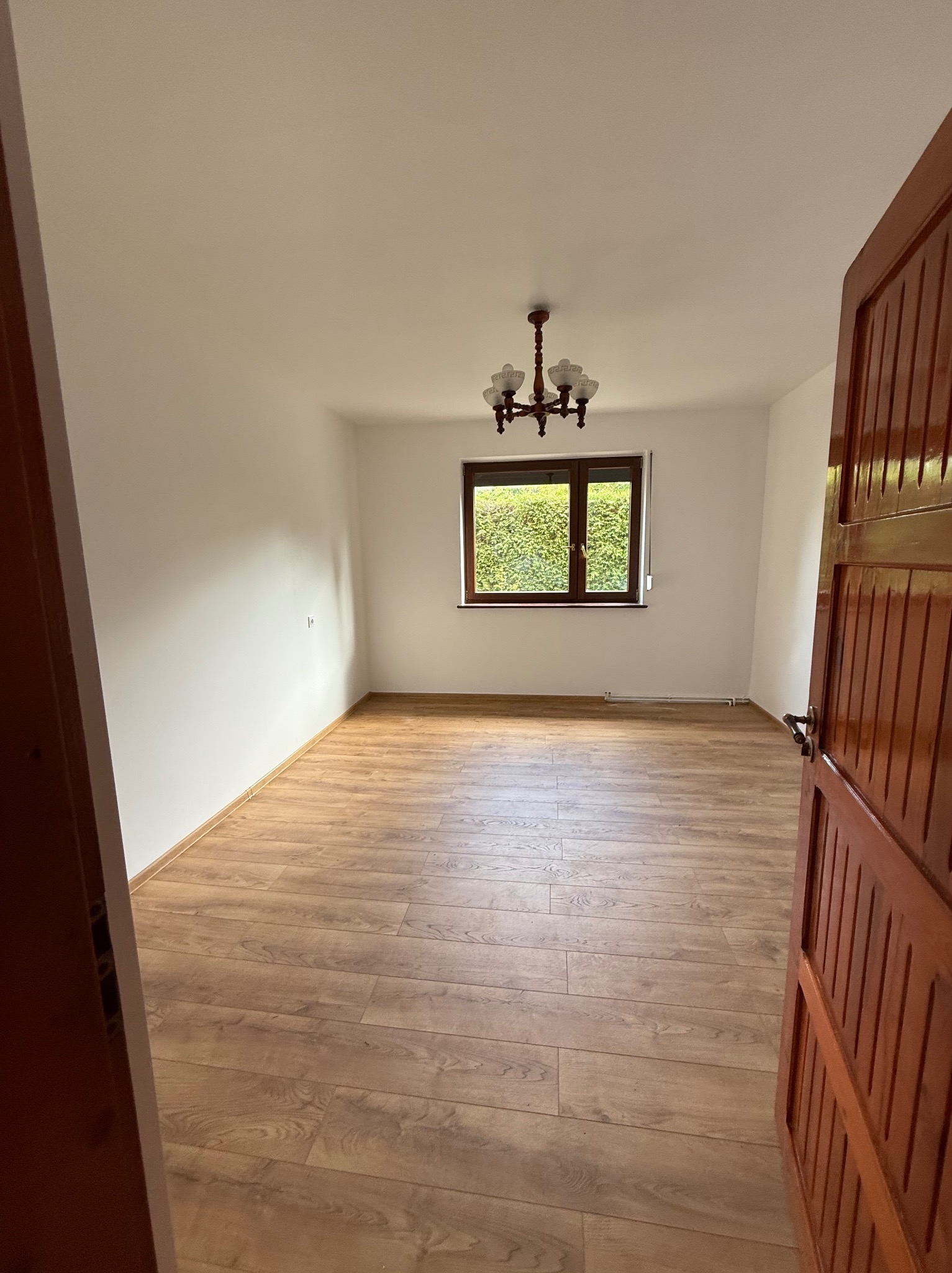 Jasne wnętrze pokoju z drewnianą podłogą, widok zza otwartych drzwi. Okno z widokiem na zieleń, klasyczny żyrandol. Świeżo wykończone ściany.