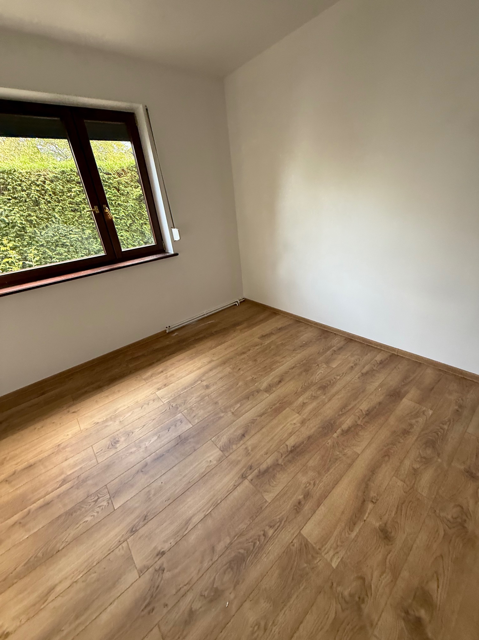 Pusty pokój po remoncie: drewnopodobna podłoga, białe ściany, okno z widokiem na zieleń. Świeżo wykończone wnętrze z widocznymi rurkami grzewczymi przy podłodze.