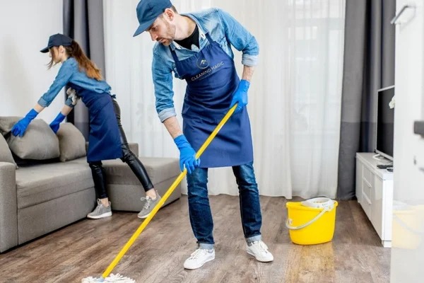Dwoje pracowników w niebieskich uniformach i rękawicach podczas mycia podłogi mopem i odkurzania sofy w jasnym salonie z drewnianą podłogą, widoczny telewizor i wiadro.