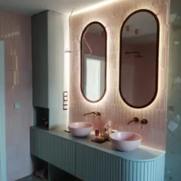 Nowoczesna łazienka z różowymi płytkami i owalnymi lustrami z podświetleniem LED. Umywalki w kolorze pudrowego różu. Szafka pod umywalką z ryflowanym frontem.