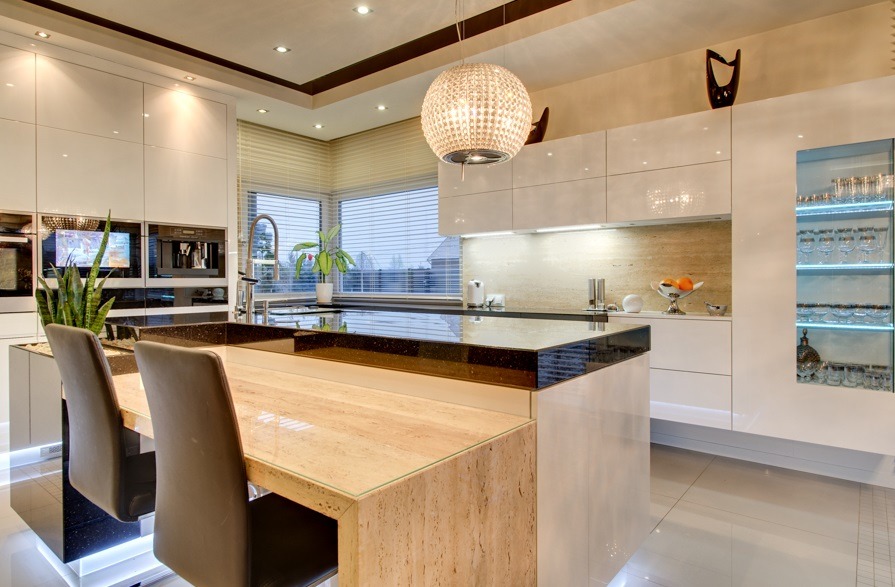 Biała, nowoczesna kuchnia z wyspą i marmurowym blatem, oświetlona lampą w kształcie kuli. Widoczne zabudowane sprzęty i szklana witryna z podświetleniem.
