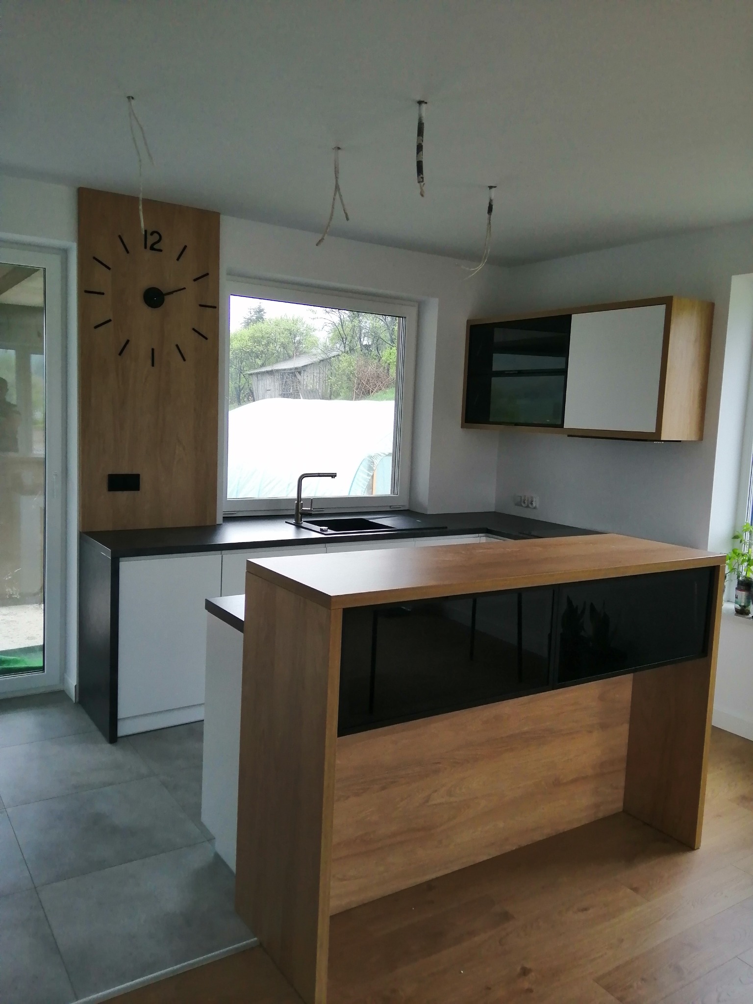 Nowoczesna kuchnia z wyspą, drewniane akcenty, minimalistyczny design, czarne blaty, wiszące kable, zegar na ścianie. Widok na zewnątrz przez okno.