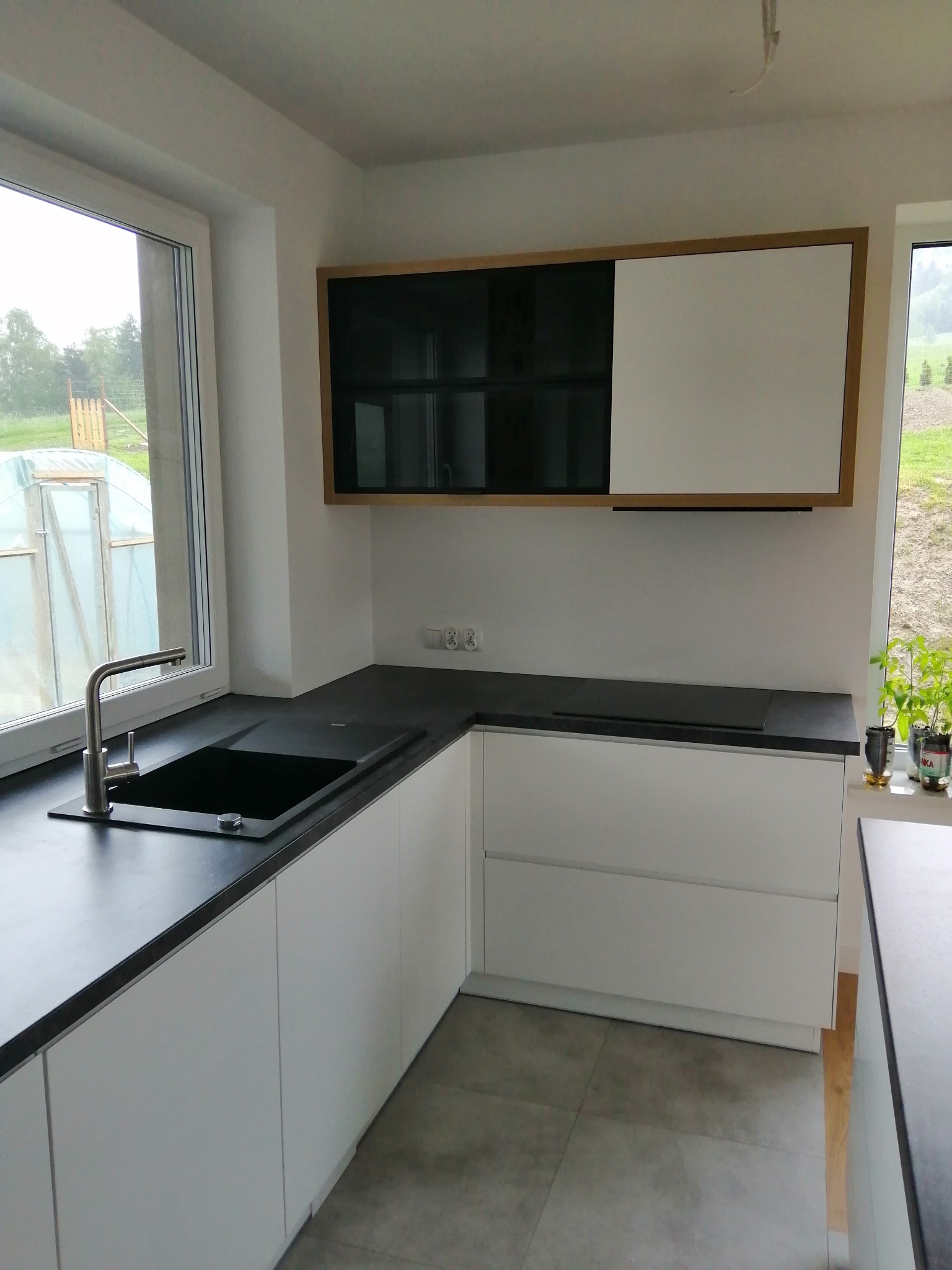 Nowoczesna kuchnia z białymi frontami i czarnym blatem, minimalistyczny design, okno z widokiem na zieleń, szafka górna z przesuwnymi drzwiami.