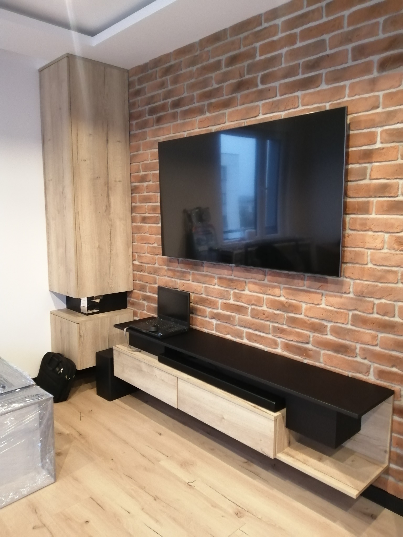 Nowoczesna szafka RTV z drewnianymi frontami i czarnym blatem, zawieszona na ceglanej ścianie. Obok wysoki regał w tym samym stylu. Na blacie laptop.
