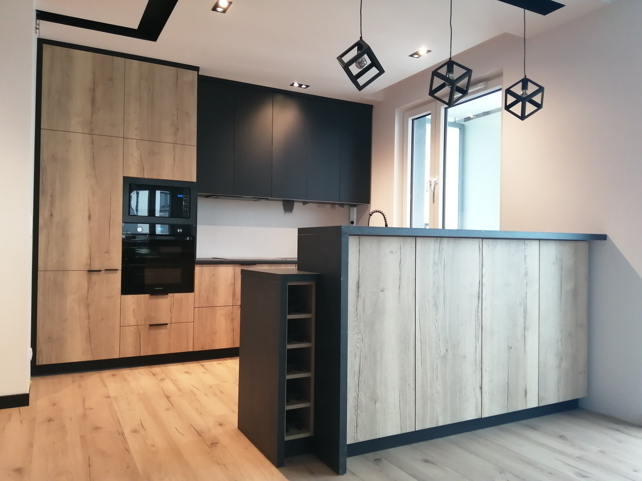 Nowoczesna kuchnia: drewniane fronty szafek, czarne elementy, wyspa z regałem na wino, geometryczne lampy wiszące. Jasne drewno na podłodze.