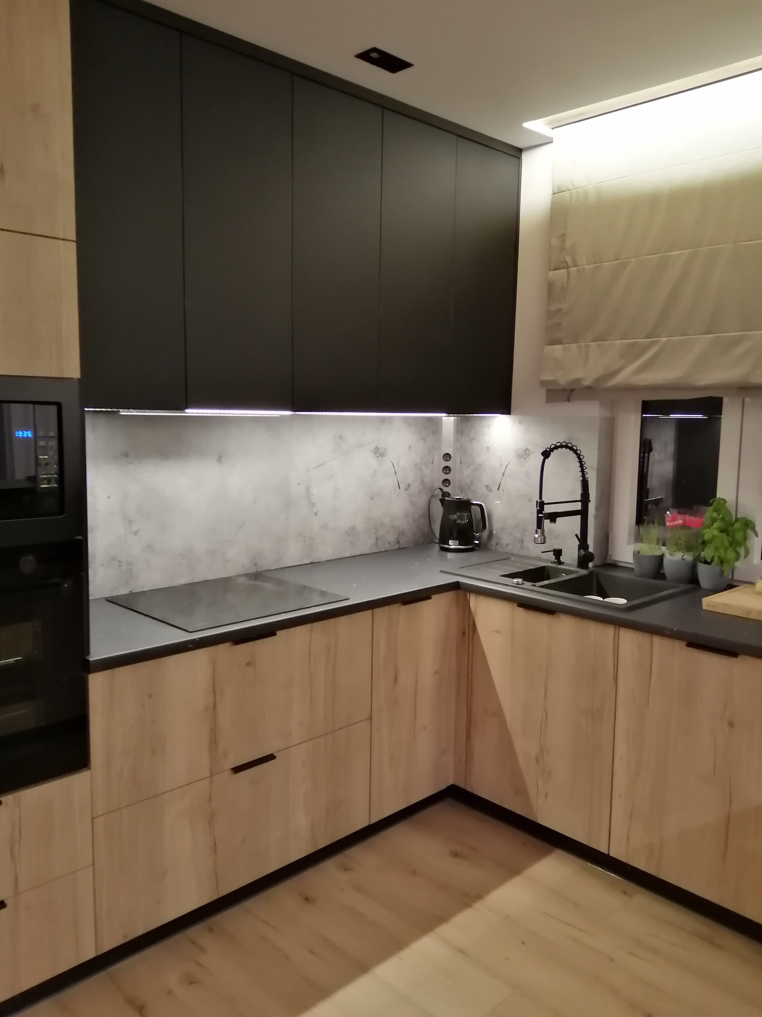 Nowoczesna kuchnia z drewnianymi szafkami dolnymi i czarnymi górnymi. Blat szary, zlew i bateria czarne. Widoczne okno z roletą rzymską i zioła w doniczkach.