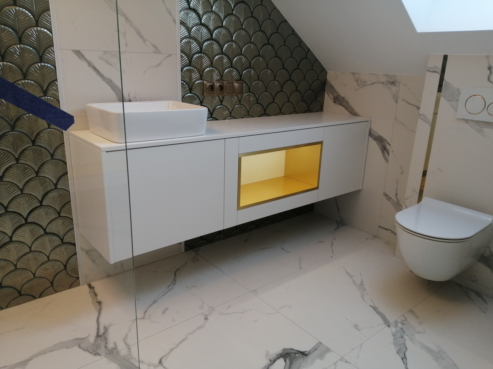 Nowoczesna łazienka na poddaszu z białą szafką pod umywalkę, złotymi akcentami i marmurową podłogą. Uwagę przykuwają płytki w kształcie wachlarzy.