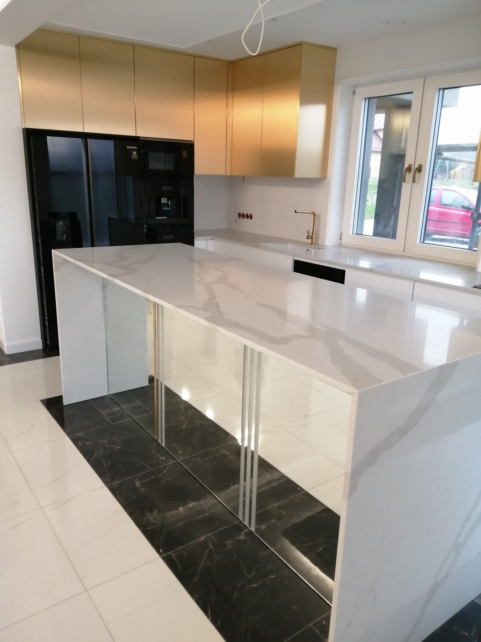 Nowoczesna kuchnia z wyspą i złotymi szafkami w Nowym Sączu. Biały blat z marmurowym wzorem, czarne AGD w zabudowie i okno z widokiem na zewnątrz.