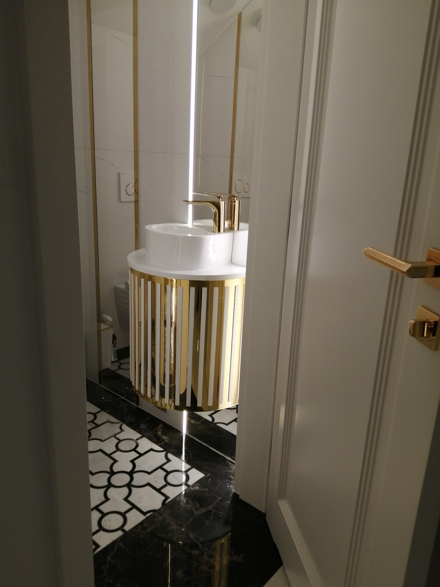 Nowoczesna łazienka z umywalką na szafce w złote pionowe paski, widoczna przez uchylone białe drzwi z pozłacaną klamką, czarno-biała podłoga.