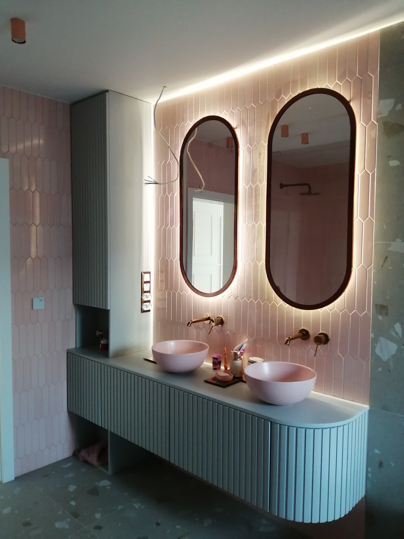 Nowoczesna łazienka z różowymi płytkami i owalnymi lustrami z podświetleniem LED. Umywalki w kolorze pudrowego różu. Szafka pod umywalką z ryflowanym frontem.