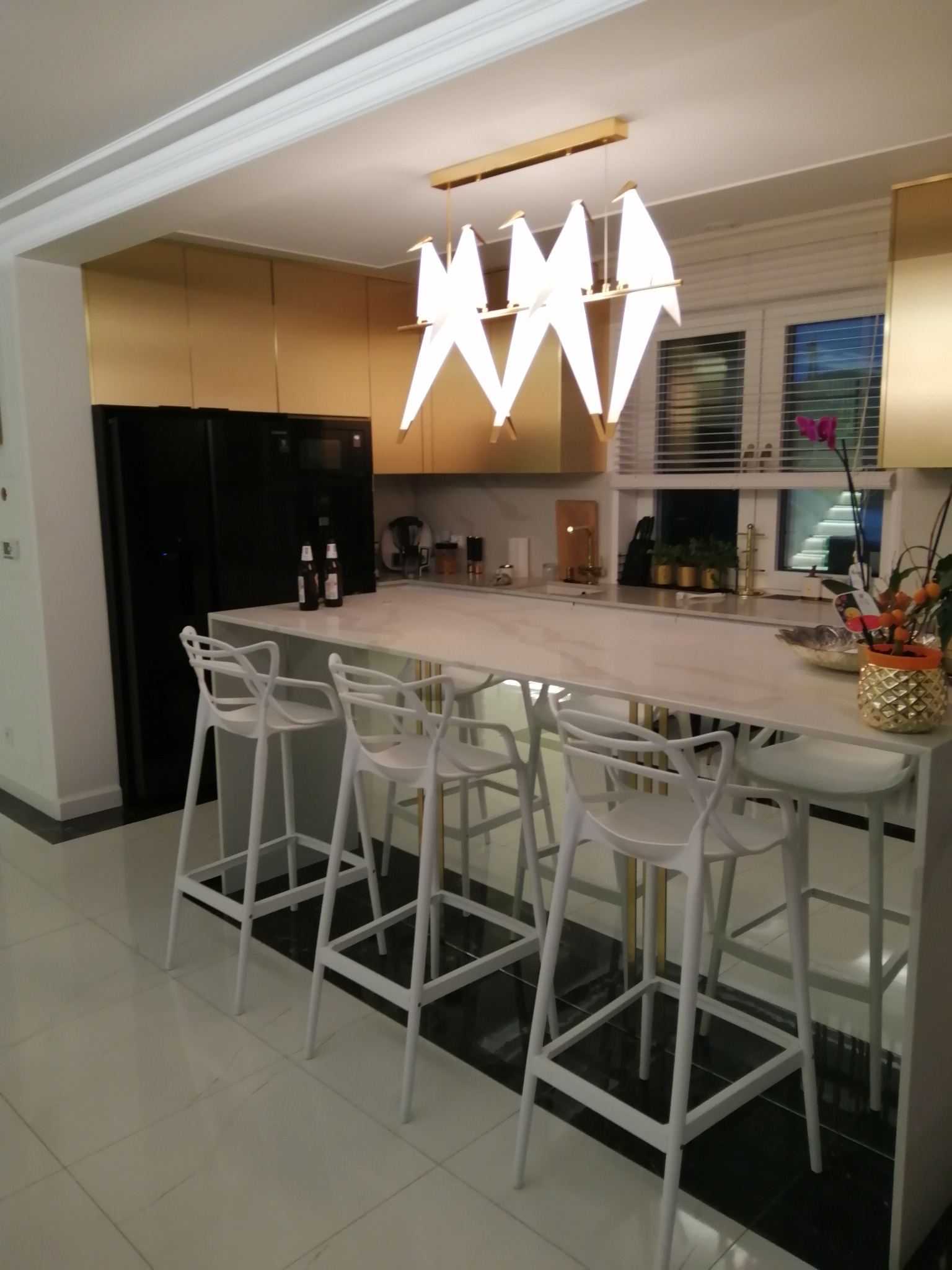 Nowoczesna kuchnia z marmurowym blatem i złotymi akcentami. Nad wyspą designerska lampa. Minimalistyczne białe krzesła barowe i czarna lodówka.