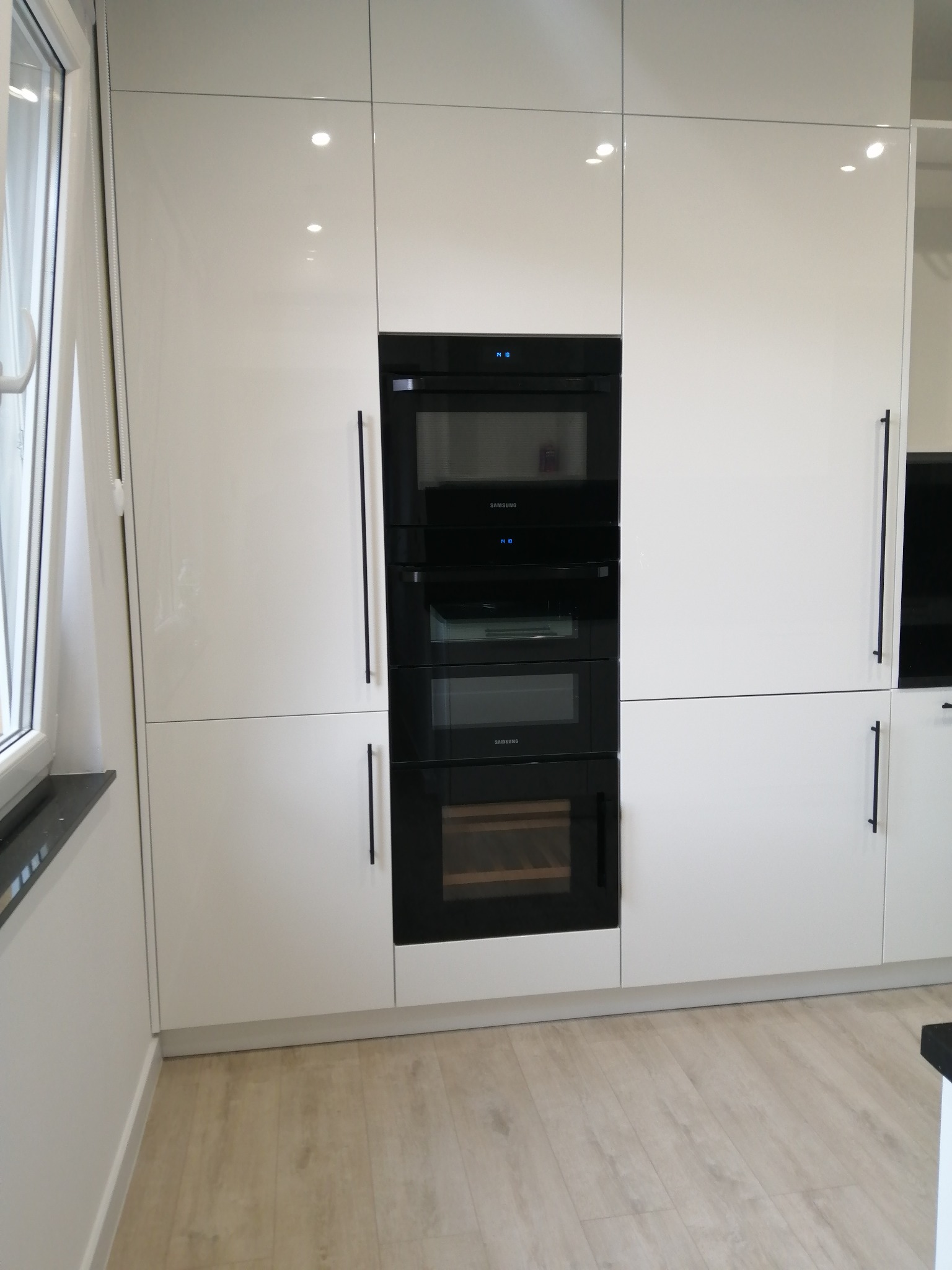 Biała, nowoczesna zabudowa kuchenna z wbudowanym sprzętem AGD (piekarnik, mikrofalówka, chłodziarka do wina) i czarnymi, długimi uchwytami, widok z perspektywy.
