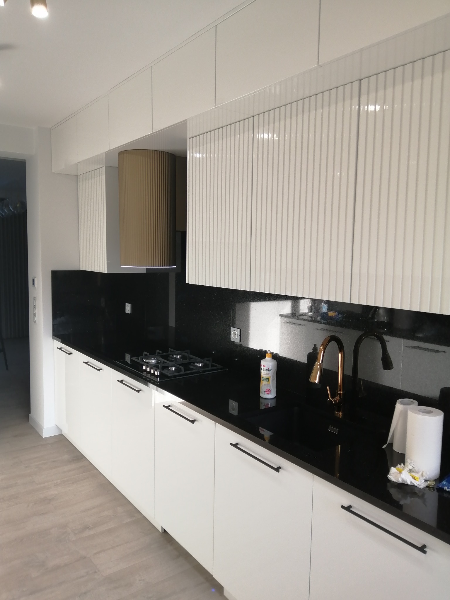 Nowoczesna kuchnia: białe, ryflowane szafki górne, czarny blat i złota armatura. Uwagę przykuwa cylindryczny okap w kolorze złota. Minimalistyczny design.