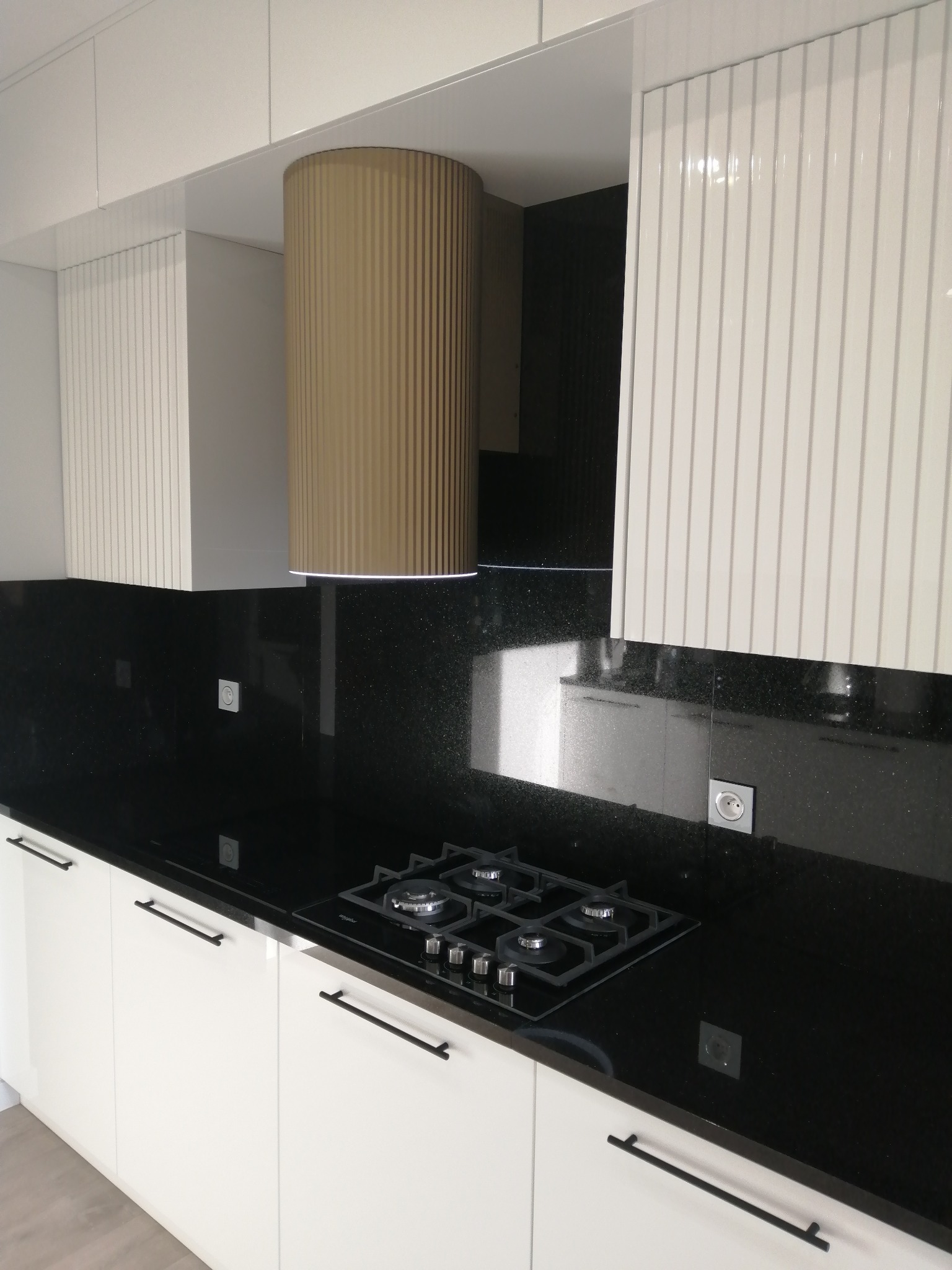 Biała kuchnia z czarnym blatem i złotym okapem. Fronty szafek z pionowymi żłobieniami. Płyta gazowa wkomponowana w blat. Minimalistyczny design.
