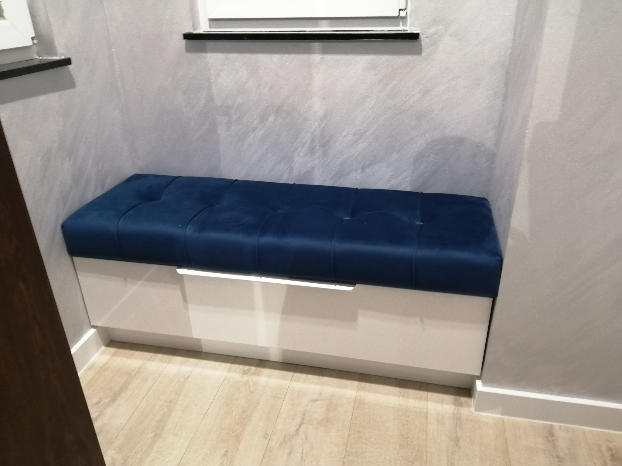 Narożna ławka tapicerowana granatowym welurem, z białym, lakierowanym frontem szuflady, wkomponowana we wnękę, na tle ściany o fakturze betonu.