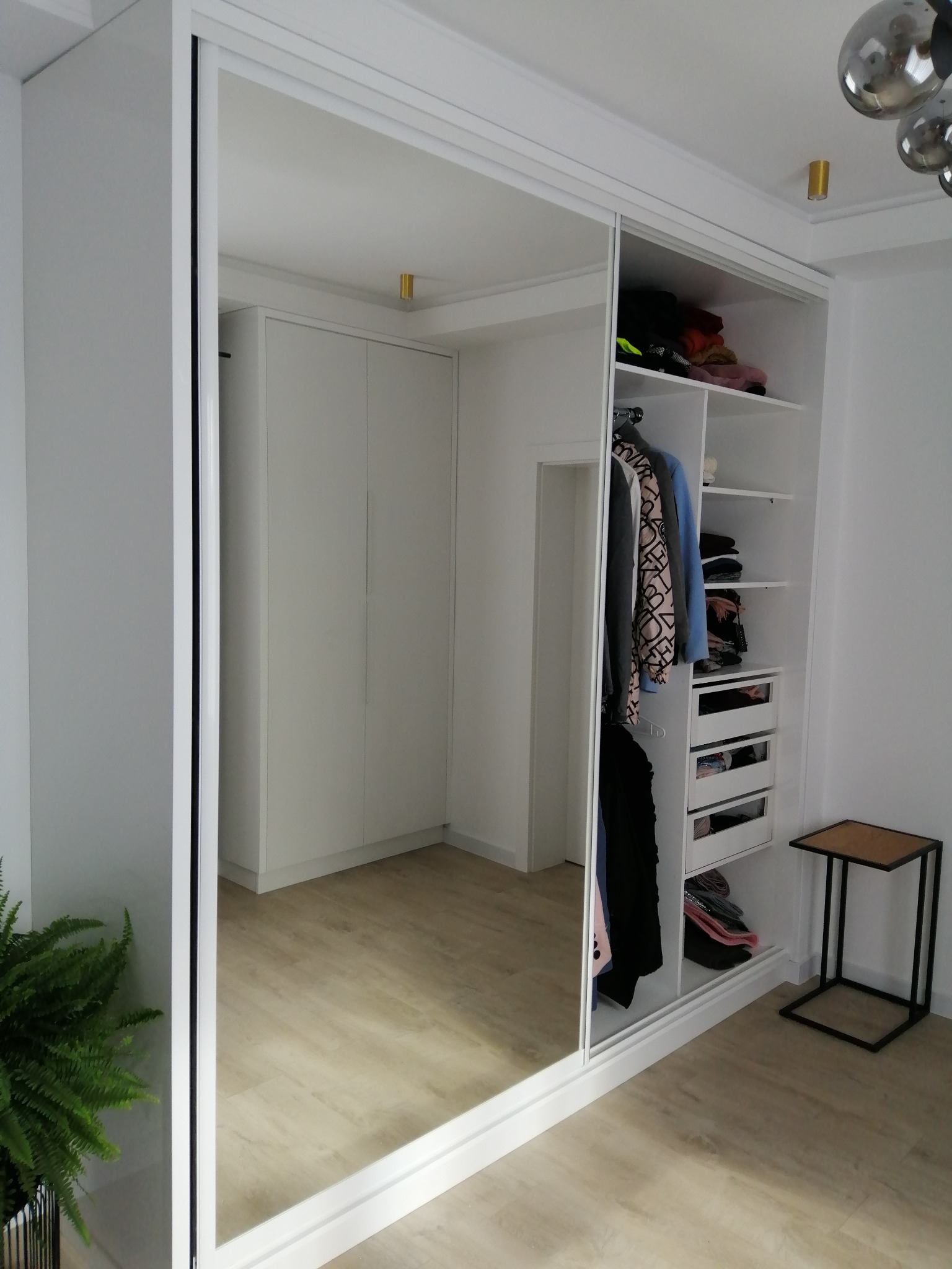 Biała szafa wnękowa z lustrzanymi drzwiami przesuwnymi, częściowo otwarta, ukazująca półki i szuflady z ubraniami. Widoczne oświetlenie punktowe i roślina doniczkowa.