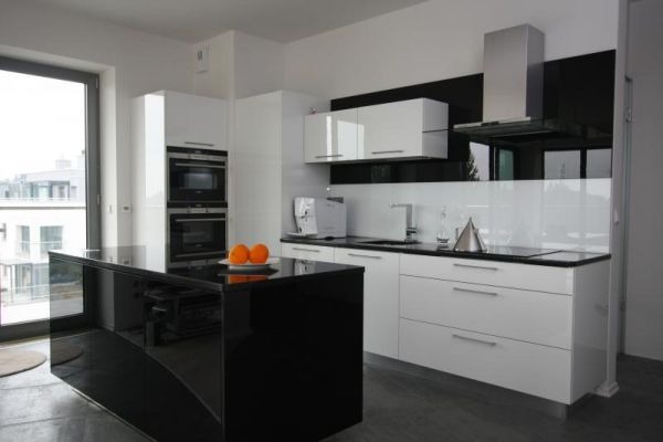 Nowoczesna kuchnia z czarnym blatem wyspowym i białymi szafkami, uzupełniona o srebrne akcenty i okno z widokiem na miasto. Minimalistyczny design.