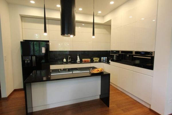 Nowoczesna kuchnia w bieli i czerni z wyspą, minimalistyczne lampy wiszące, zabudowane sprzęty AGD. Parkiet na podłodze.