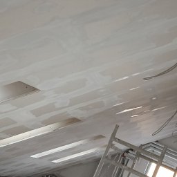 MAR-BUD Mateusz Potyński - Szpachlowana sufitowa płyta G-K z widocznymi otworami na oświetlenie, częściowo wykończona. W tle drabina i instalacje. Ujęcie z dołu pod kątem.