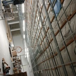 MAR-BUD Mateusz Potyński - Montaż metalowej konstrukcji pod płyty na ścianie, widoczne ocieplenie i instalacje. Pracownik na rusztowaniu. Ujęcie z dołu, perspektywa podkreśla wysokość.