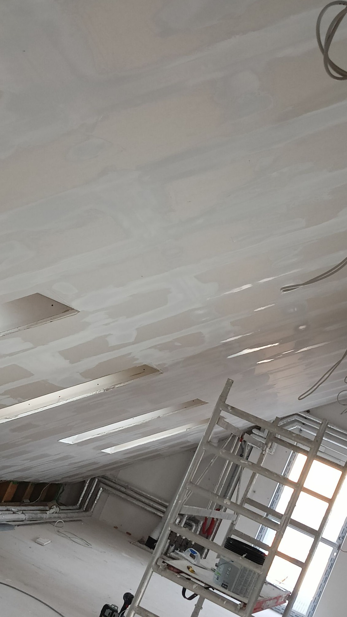 Szpachlowana sufitowa płyta G-K z widocznymi otworami na oświetlenie, częściowo wykończona. W tle drabina i instalacje. Ujęcie z dołu pod kątem.