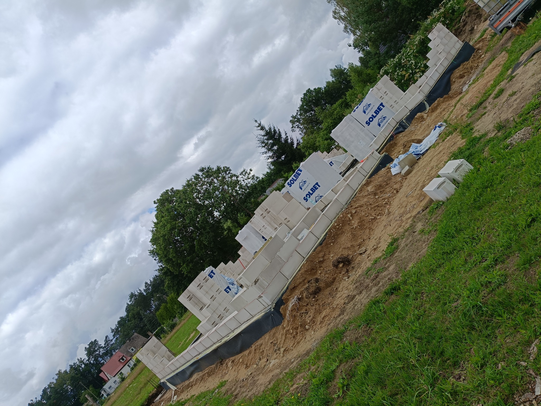Budowa fundamentów z bloczków Solbet na działce z zielenią, widok pod kątem. Częściowo wzniesione ściany, pakowane bloczki czekają na montaż.