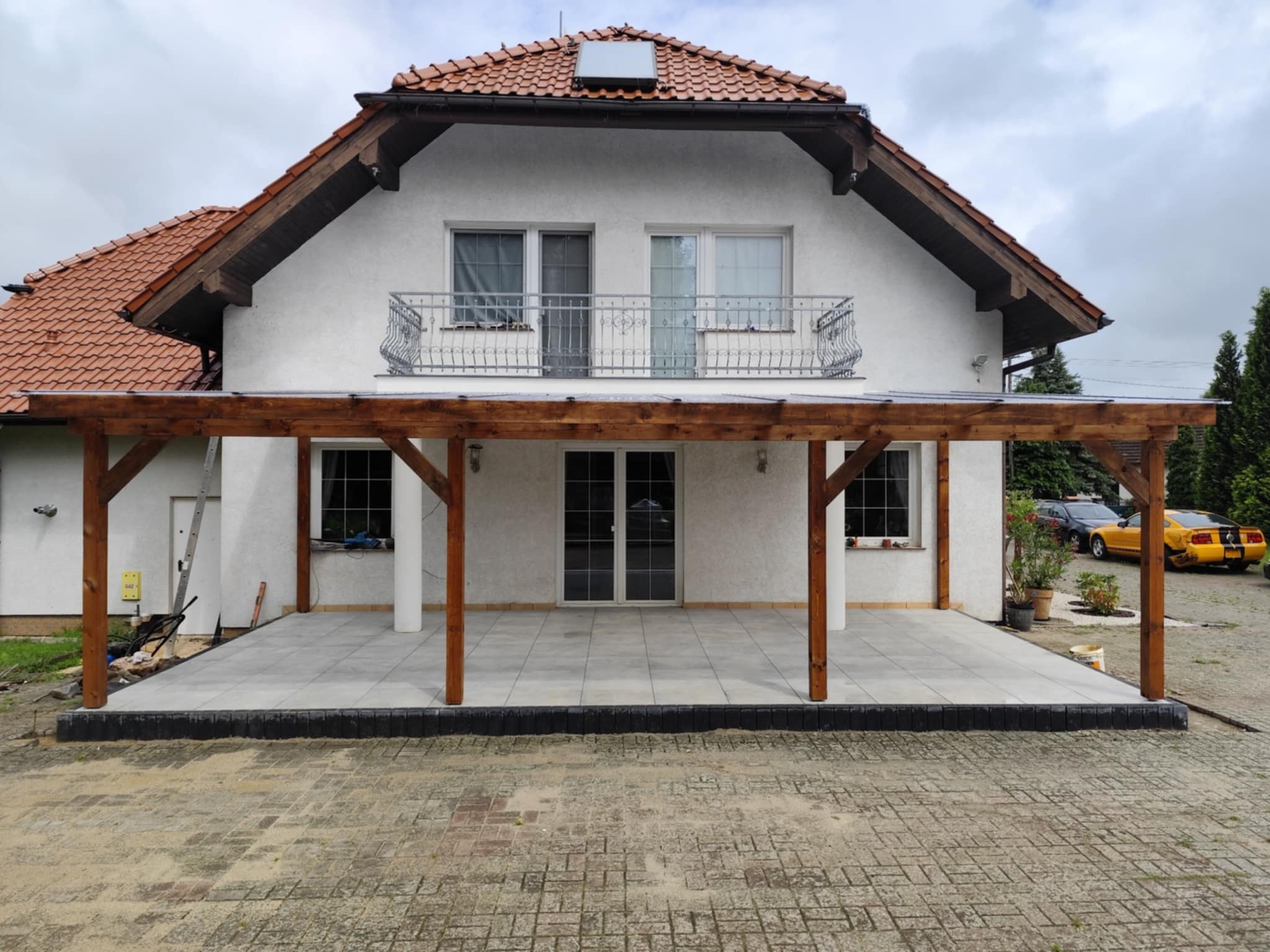 Nowoczesny taras z drewnianą pergolą przy białym domu z balkonem. Ułożona szara kostka, widoczne czarne obrzeża, elementy wykończeniowe. W tle zaparkowany żółty samochód.