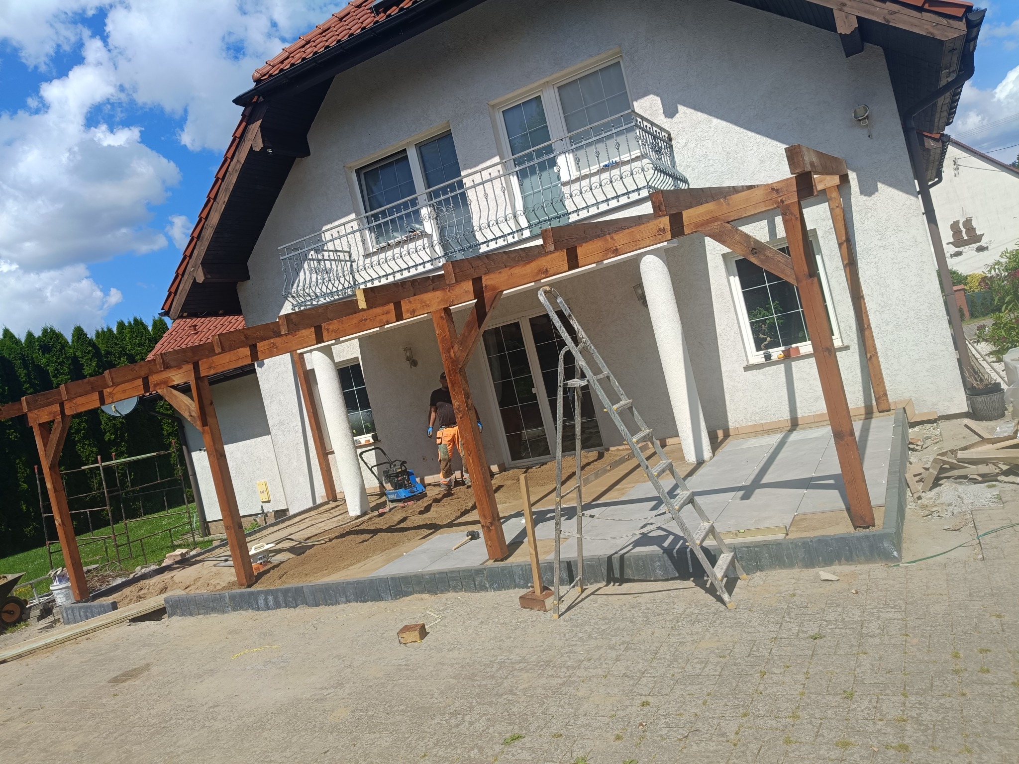 Drewniana pergola przy domu w trakcie budowy, widoczne kolumny, częściowo ułożona kostka brukowa, narzędzia i pracownik w oddali. Słoneczny dzień.
