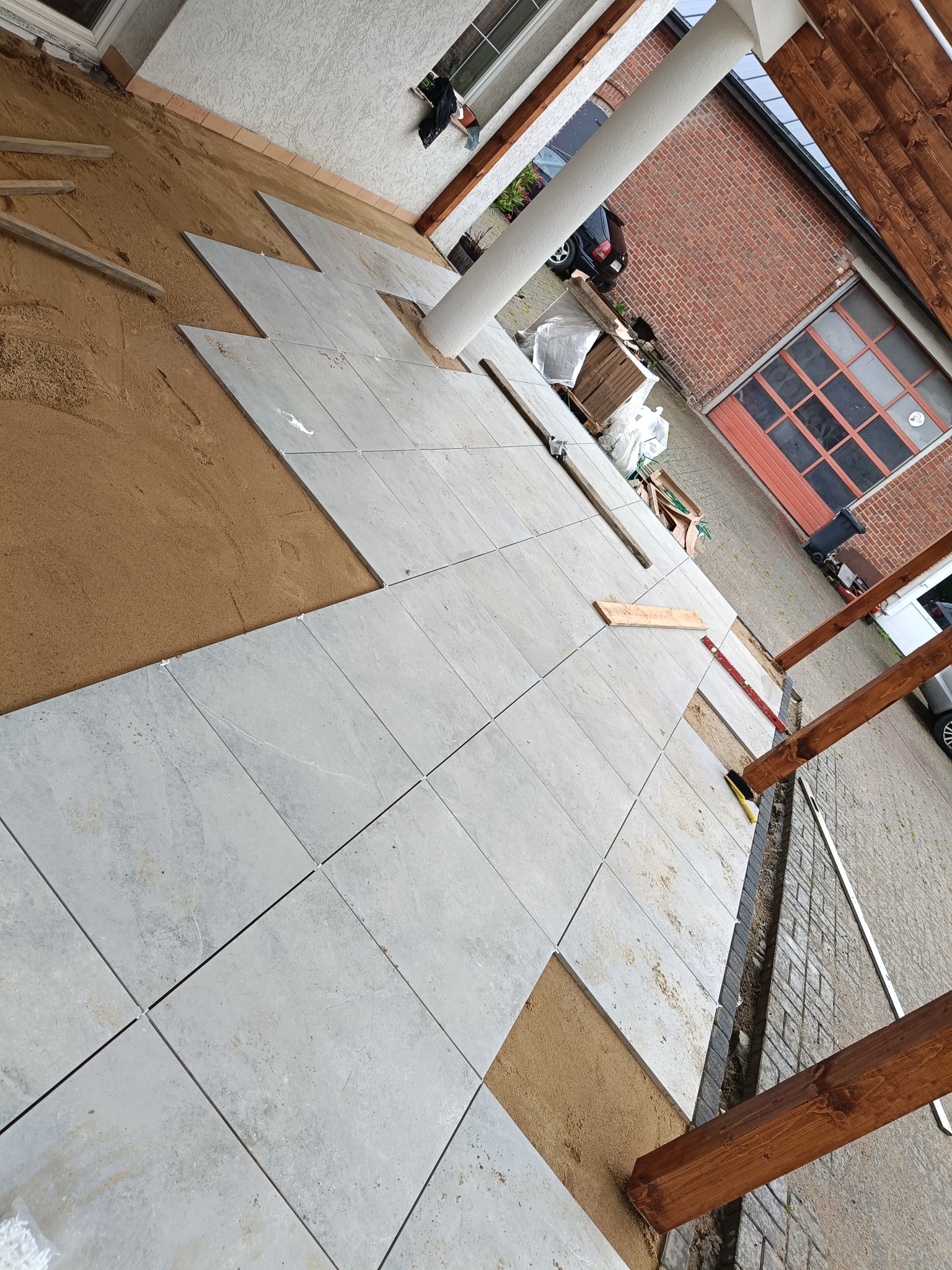 Układanie jasnych, kwadratowych płytek na piasku, widoczne drewniane elementy konstrukcji zadaszenia. Częściowo wykończona powierzchnia.