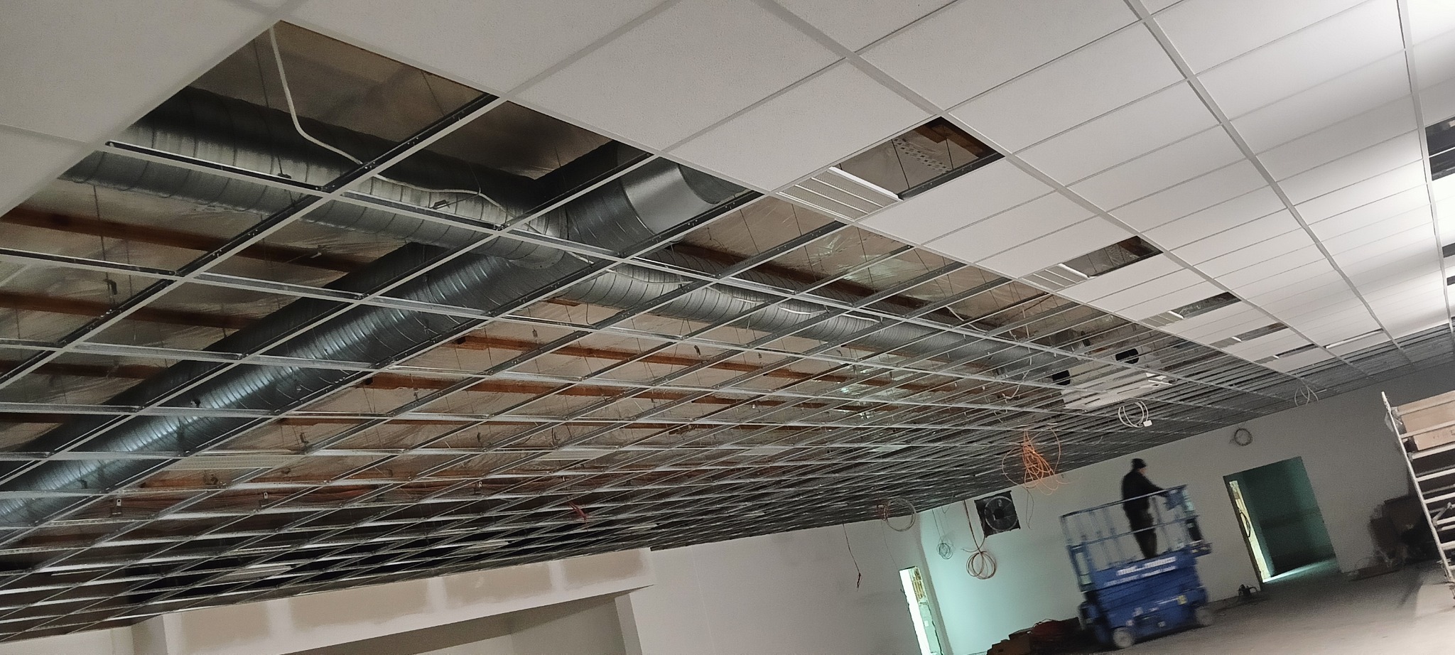 Montaż sufitu podwieszanego kasetonowego z widoczną konstrukcją i instalacją wentylacyjną. Pracownik na podnośniku koszowym w tle.