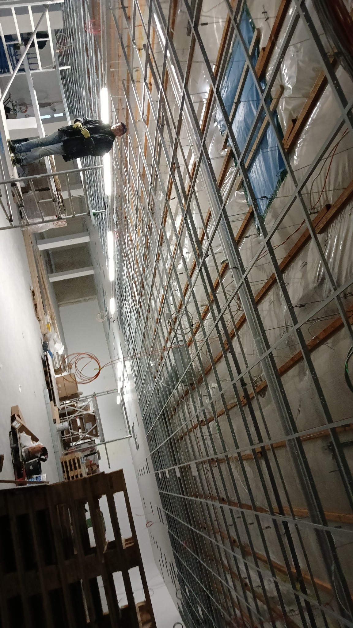 Montaż metalowej konstrukcji pod płyty na ścianie, widoczne ocieplenie i instalacje. Pracownik na rusztowaniu. Ujęcie z dołu, perspektywa podkreśla wysokość.