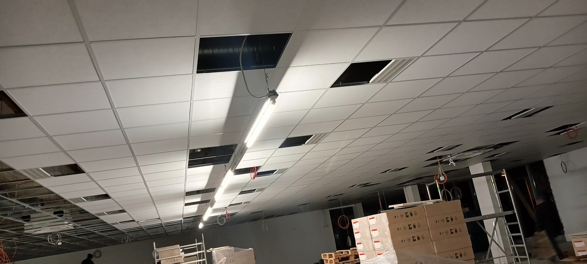 Montaż sufitu podwieszanego z widocznymi elementami instalacji i oświetleniem. Część paneli brakuje, ukazując konstrukcję nośną.